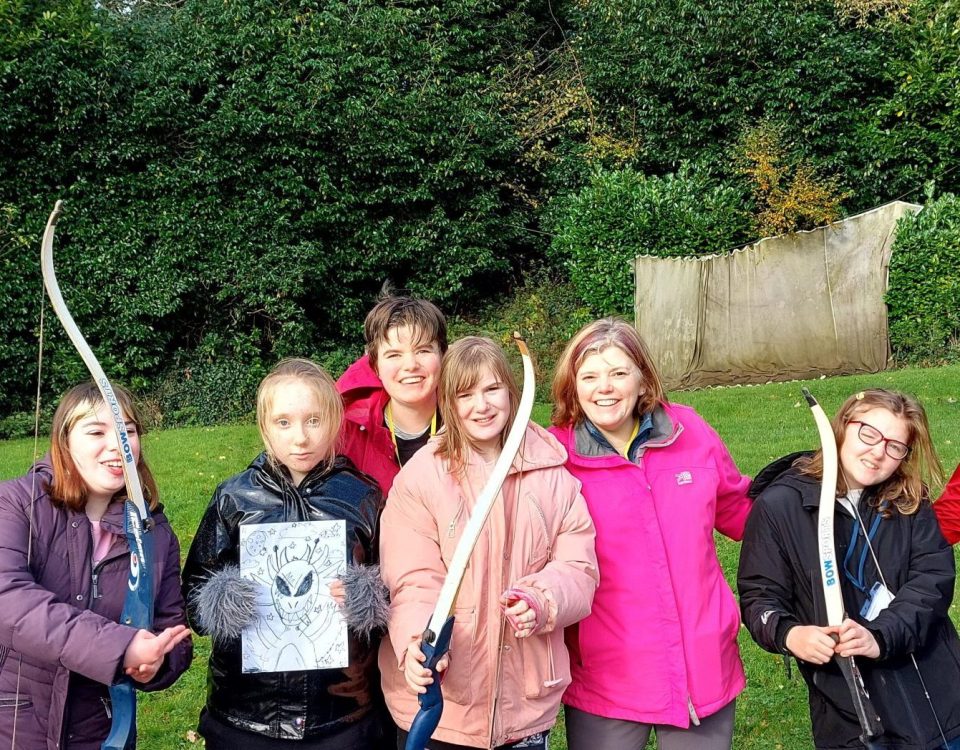 A group of young people and leaders on the Additional Support Needs weekend standing in front of the grass archery range, smiling at the camera and holding archery bows.